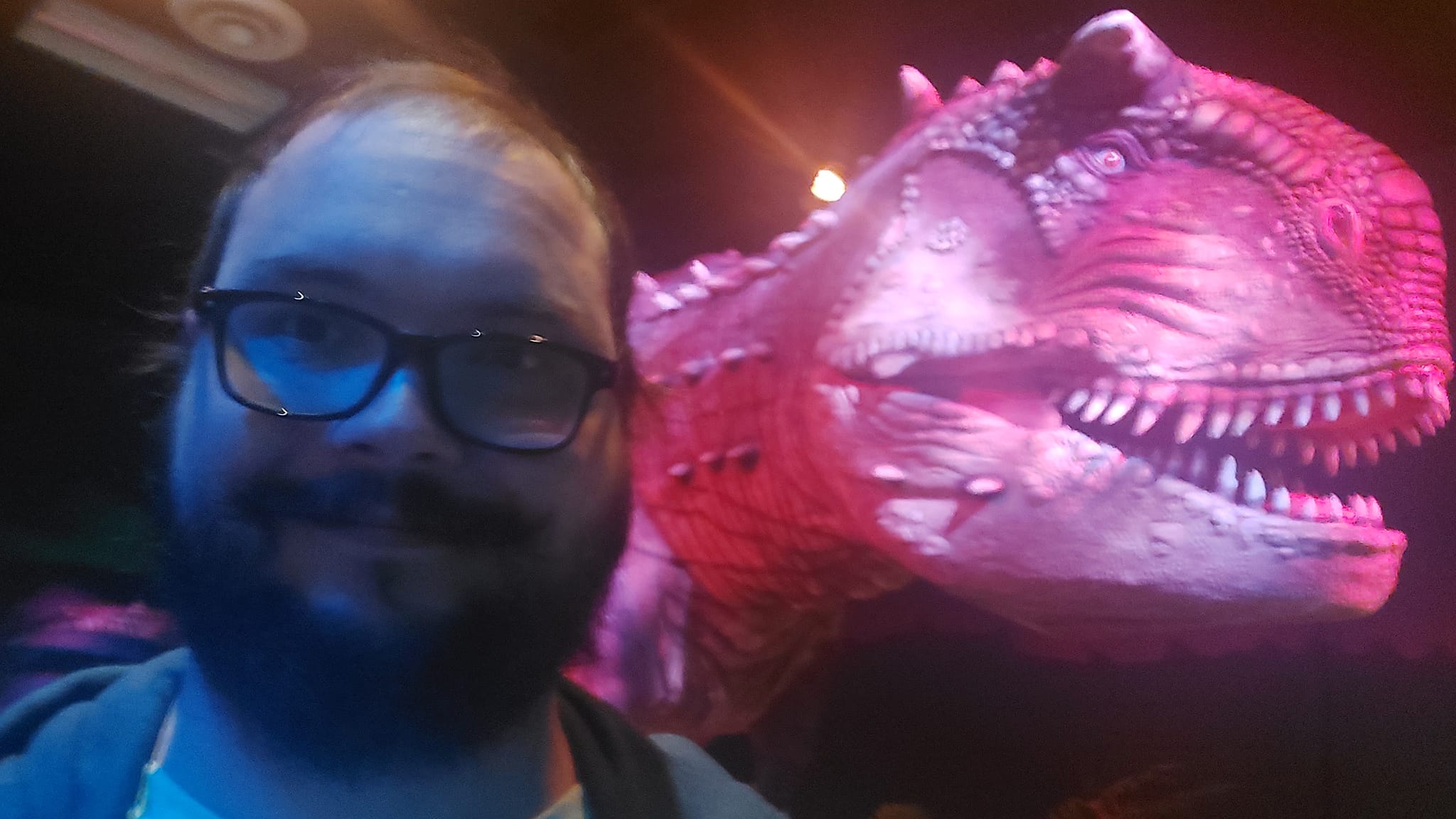 A man with a ball cap and bushy facial hair is standing in front of a carnotaurus animatronic